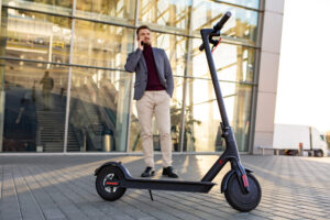 Home - young handsome man with e scooter standing sidewalk near airport sunset talking smartphone 1 scaled